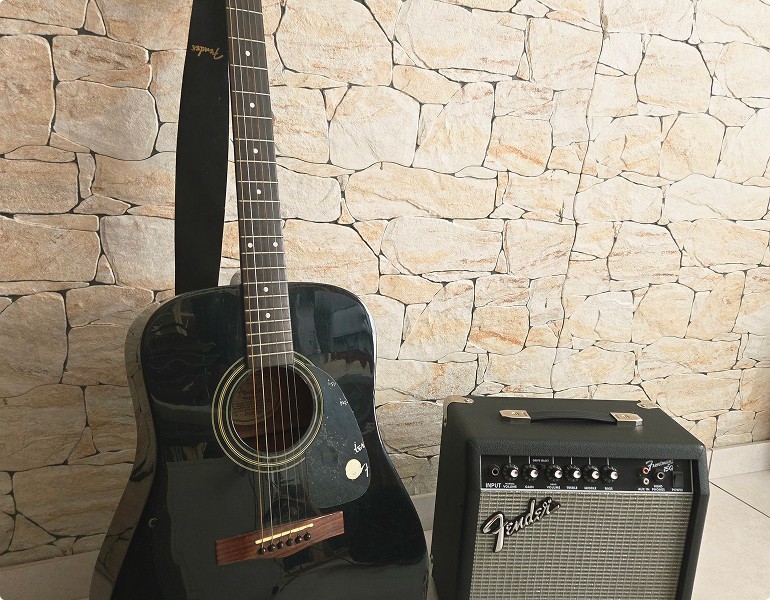 Guitare acoustique noire et amplificateur Fender placés contre un mur à motif de pierre.